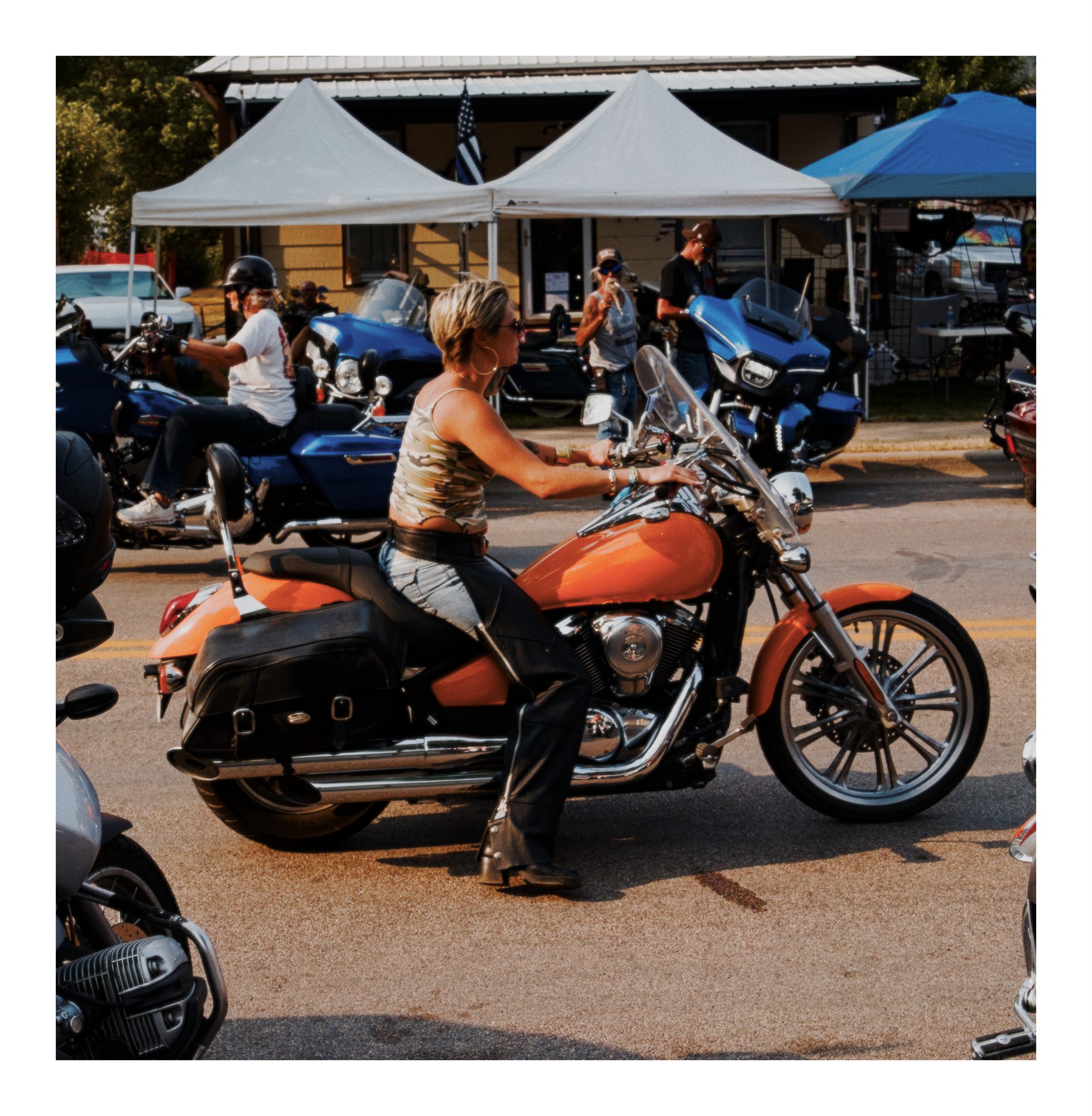 A rider seated on an orange motorcycle near tents and parked bikes.